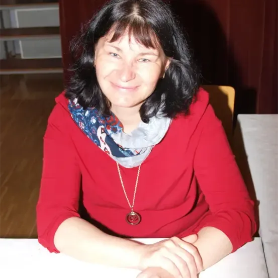 Eine Frau in einem roten Shirt und Schal sitzt lächelnd am Tisch, mit einer Treppe und einer roten Wand im Hintergrund.