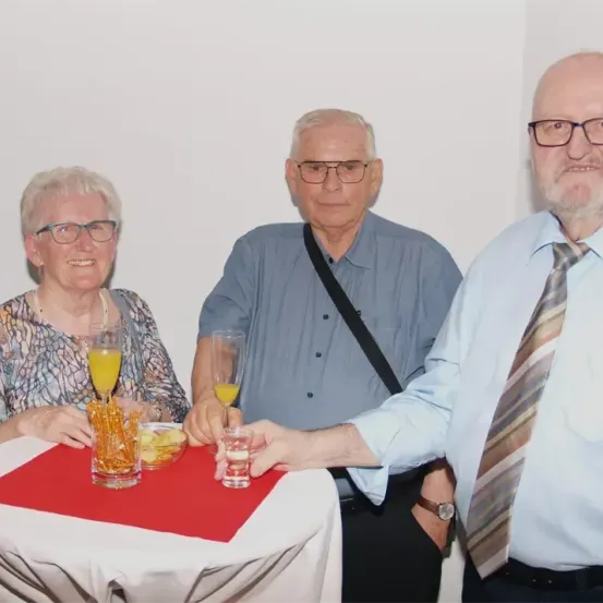 Drei ältere Menschen, zwei Männer und eine Frau, stehen an einem Tisch mit Getränken. Die Frau trägt eine Brille und eine Halskette. Die Männer tragen Krawatten und Brillen. Sie schauen in die Kamera.