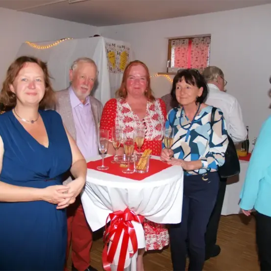 Vier Erwachsene stehen um einen Tisch herum, lächeln und halten Weingläser. Eine Frau in einem blauen Kleid und ein Mann in einem rosa Hemd stehen links. Eine weitere Frau in einem Blumenkleid und eine Frau in einem blau-weiß gemusterten Shirt stehen rechts. Dahinter befindet sich eine weiße Gardine mit Dekorationen.