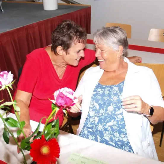 Zwei Frauen sitzen an einem Tisch, lächeln, wobei eine Frau eine rosa Blume hält und die andere Frau eine weiße Jacke trägt.