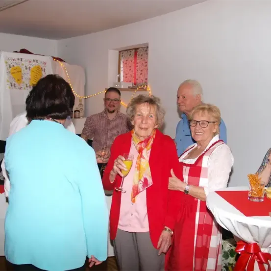 Eine Gruppe von Menschen, darunter eine ältere Frau in einem roten Mantel, steht und unterhält sich in einem Raum. Hinter ihnen steht ein Mann in einem blauen Hemd in der Nähe eines Tisches mit Weingläsern.