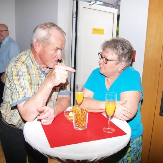 Ein älteres Paar, das an einem Tisch sitzt und Orangensaft genießt. Der Mann deutet mit einem leichten Lächeln auf etwas, während die Frau aufmerksam zuhört. Hinter ihnen steht eine Tür mit der Aufschrift 'Entrée' und eine verschwommene Figur.