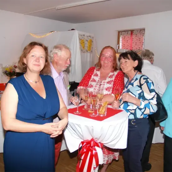Eine Gruppe von Menschen bei einem Event, zwei Frauen vorne mit Champagnergläsern, ein Mann dahinter und andere im Hintergrund.