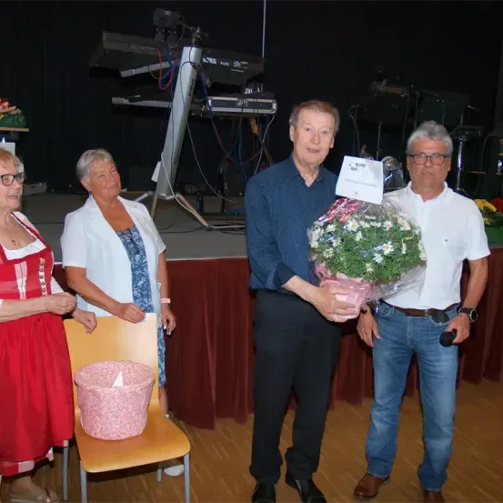 Vier Personen stehen auf einer Bühne mit Musikequipment im Hintergrund. Ein älterer Mann hält einen Blumenstrauß. Eine Frau in einem roten Kleid lächelt neben einem Stuhl mit einem rosa Korb.
