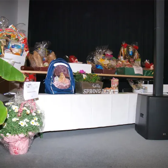 Ein Tisch mit weißer Tischdecke zeigt verschiedene Geschenkkörbe und einen blauen Rucksack, zusammen mit einer Blumendekoration. Dahinter befindet sich ein Holzregal mit weiteren Geschenkkörben. Ein Schild sagt 'Hallo Frühling' und ein Lautsprecher steht rechts.