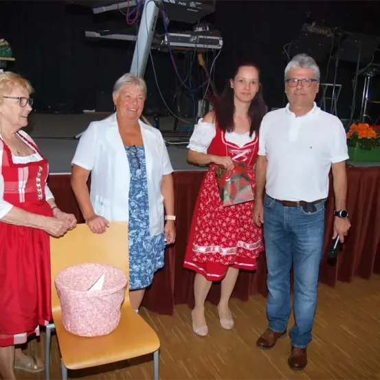 Vier Personen stehen auf einer Bühne. Eine Frau in einem roten Kleid hält ein Geschenk. Eine ältere Frau in einem roten Kleid und eine andere in einer weißen Jacke stehen neben ihr. Ein Mann in einem weißen Hemd und blauen Jeans steht rechts.