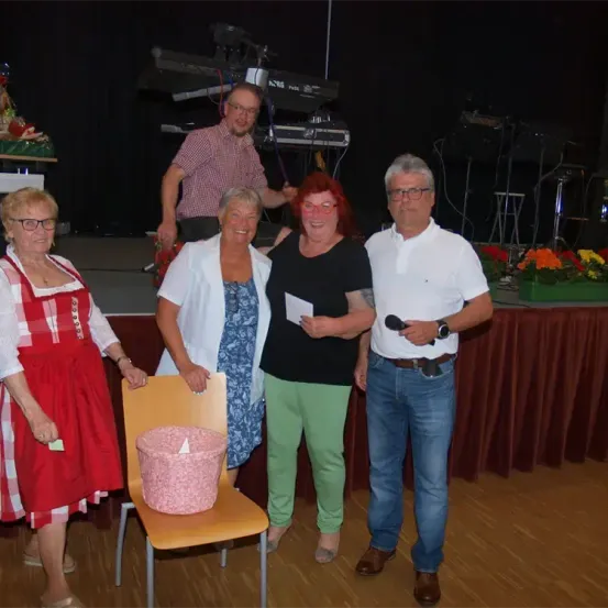 Fünf Erwachsene stehen auf einer Bühne, mit einer Person, die Klavier spielt, im Hintergrund. Eine Frau sitzt auf einem Stuhl mit einem rosa Eimer darauf.