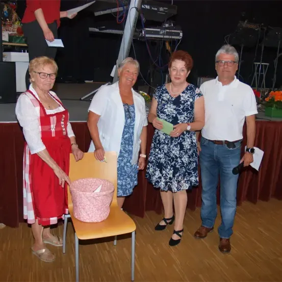 Vier Personen, darunter zwei Frauen in Kleidern und ein älterer Mann in einem weißen Hemd, stehen gemeinsam auf einer Bühne mit Musikinstrumenten im Hintergrund.