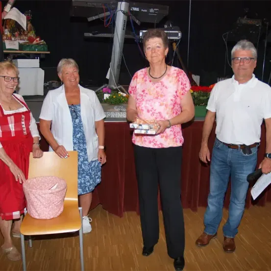 Vier Personen stehen in einem Raum, wahrscheinlich bei einer Veranstaltung. Eine Frau in der Mitte hält ein Geschenk. Die Frauen tragen Kleider und die Männer Brillen und einen Gürtel. Hinter ihnen steht ein Tisch mit einer Topfpflanze.