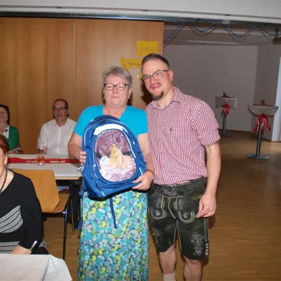 Eine Frau und ein Mann stehen in einem Raum, lächeln und halten einen Rucksack. Sie sind in Freizeitkleidung gekleidet. Hinter ihnen sitzen Menschen an einem Tisch. Der Raum hat Holzböden und ein gelbes Schild an der Wand.