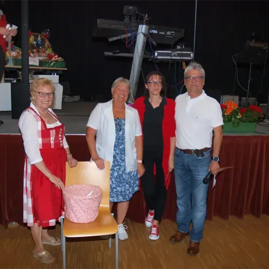 Vier Menschen stehen auf einer Bühne mit einem roten Tischtuch und lächeln für ein Foto. Die Frau links trägt ein rotes Schürzen. Ein rosa Korb sitzt auf einem Stuhl zwischen ihnen. Dahinter steht ein Tisch mit einem Korb, einem schwarzen Vorhang und Musikequipment.