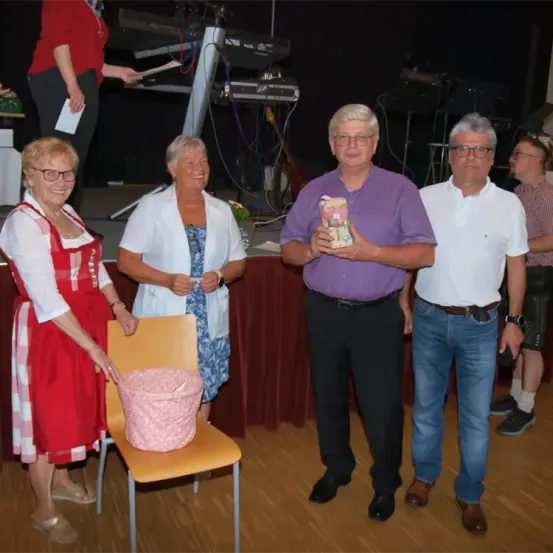 Fünf Personen stehen in einem Raum. Ein Mann in einem lila Hemd hält ein Geschenk. Eine Frau in einem roten Kleid steht neben einem Stuhl mit einem rosa Korb. Dahinter steht ein Klavier und eine weitere Person.