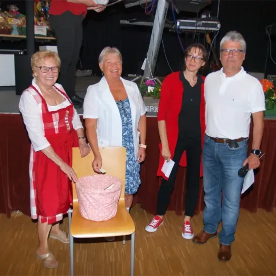 Vier Personen stehen auf einer Bühne mit einem Stuhl und einem Korb. Zwei Frauen und zwei Männer, alle lächeln. Die Frau links trägt ein rotes Kleid, die rechte eine weiße Jacke. Der Mann rechts hält ein Mikrofon.
