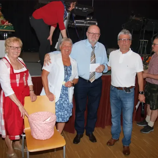 Fünf Menschen stehen auf einer Bühne. Zwei Frauen und drei Männer. Eine Frau hält eine rosa Tasche. Eine andere Frau hält ein Papier. Ein Mann hält ein Papier. Ein Mann lehnt sich gegen die Wand.