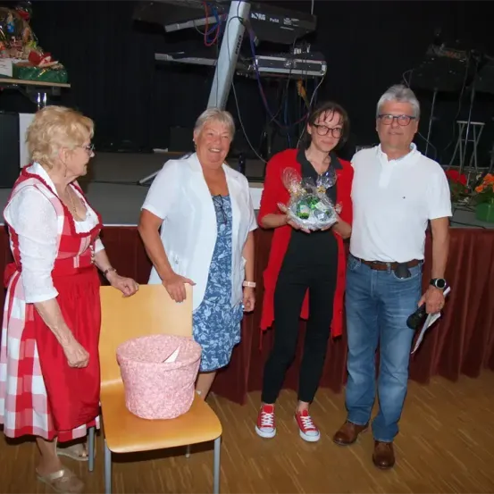 Vier Personen stehen auf einer Bühne, zwei Frauen und zwei Männer. Eine Frau in einer roten Jacke hält einen Geschenkkorb. Die Frau im weißen Kleid lächelt. Ein rosafarbener Korb sitzt auf einem Stuhl.