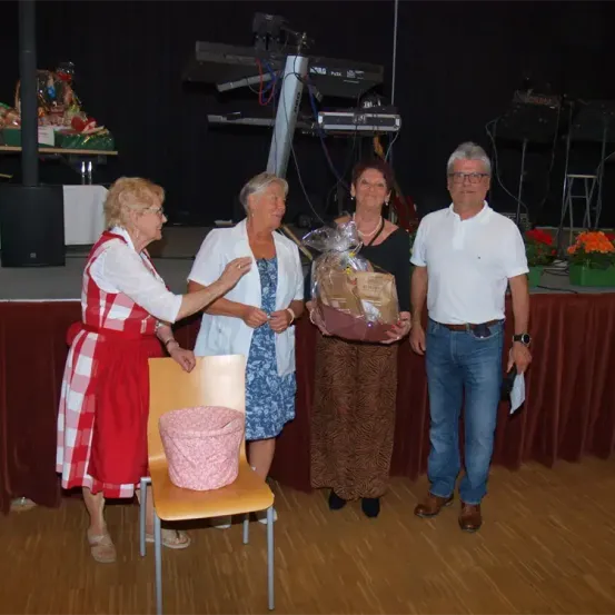 Eine Gruppe von Menschen auf einer Bühne, zwei Frauen und ein Mann, halten ein Geschenk. Die Frau links trägt ein rot-weiß kariertes Kleid.