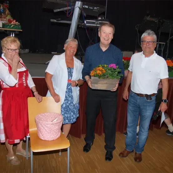 Vier Erwachsene stehen auf einer Bühne. Ein Mann in einem blauen Hemd hält eine Blumenarrangement, während eine Frau in einem roten Kleid und eine andere in einer weißen Jacke lächeln. Die Bühne hat einen Holzboden und einen roten Vorhang als Hintergrund.
