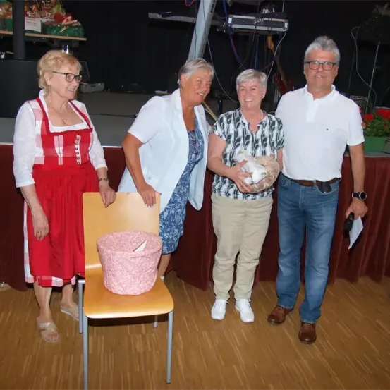 Vier Menschen stehen lächelnd auf einer Bühne. Eine Frau in einem roten Kleid, eine Frau in einer weißen Jacke, eine Frau in einem weißen und schwarzen Shirt und ein Mann in einem weißen Hemd. Ein Korb steht auf einem Stuhl.