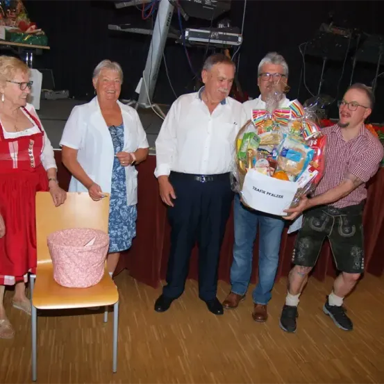 Fünf Personen stehen zusammen, wobei zwei Männer einen Geschenkkorb halten, während zwei Frauen in der Nähe stehen. Ein Stuhl mit einem rosa Behälter steht vor ihnen. Der Hintergrund zeigt eine Bühne mit Musikinstrumenten.