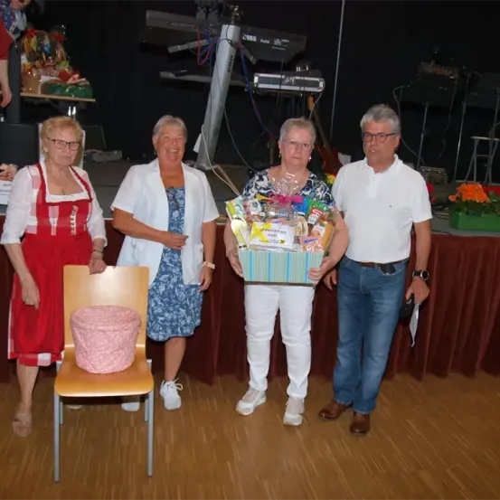 Vier Menschen posieren für ein Foto in einem Raum mit Holzboden. Eine Frau hält eine Geschenkbox. Hinter ihnen ist ein Tisch mit Blumen und ein Stuhl zu sehen.