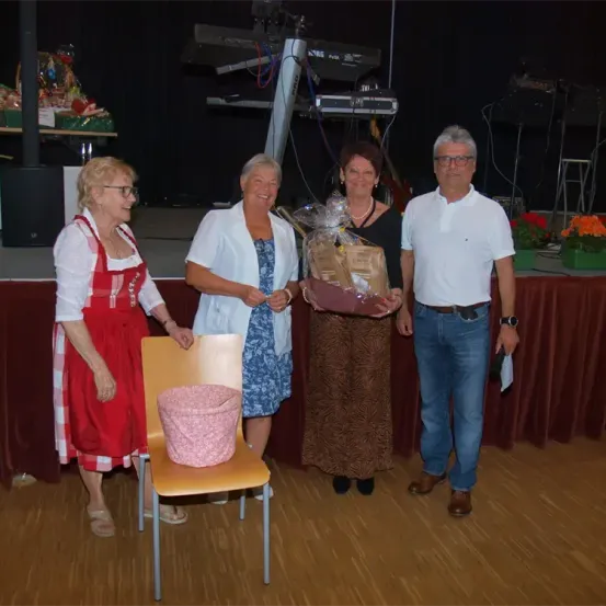 Vier Menschen stehen gemeinsam auf einer Bühne. Eine Frau hält einen Korb. Ein Stuhl steht neben einer Frau in einem roten Kleid.