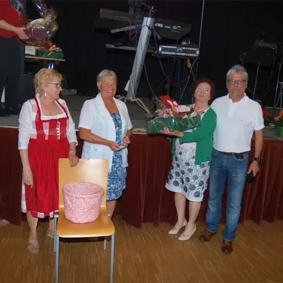 Vier Erwachsene stehen lächelnd auf einer Bühne. Eine Frau hält einen Geschenkkorb. Dahinter steht ein Tisch mit einem Geschenk und einem Klavier.
