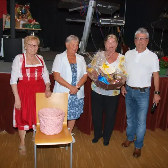 Vier Personen, drei Frauen und ein Mann, stehen vor einer Bühne. Die Frau auf der linken Seite trägt ein rotes Kleid, während die anderen weiße und blaue Kleidung tragen. Die Frau in der Mitte hält einen Geschenkkorb. Ein Stuhl mit einem rosa Bezug steht zwischen ihnen. Hinter ihnen befindet sich eine Bühne mit einem Tisch und Musikinstrumenten.