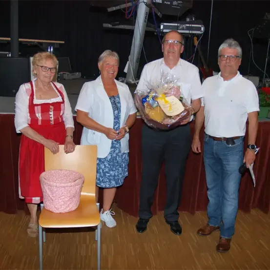 Vier Personen stehen auf einer Bühne. Ein Mann hält einen Korb mit Essen. Eine Frau in einem roten Kleid sitzt auf einem Stuhl. Eine weitere Frau steht neben ihm. Die Bühne hat einen roten Vorhang.