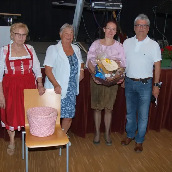 Vier Personen stehen auf einer Bühne, zwei Frauen und zwei Männer. Die Frau auf der linken Seite trägt ein traditionelles rotes und weißes Kleid, während die andere Frau einen Geschenkkorb hält. Der Mann auf der rechten Seite trägt einen Gürtel und eine Brille.
