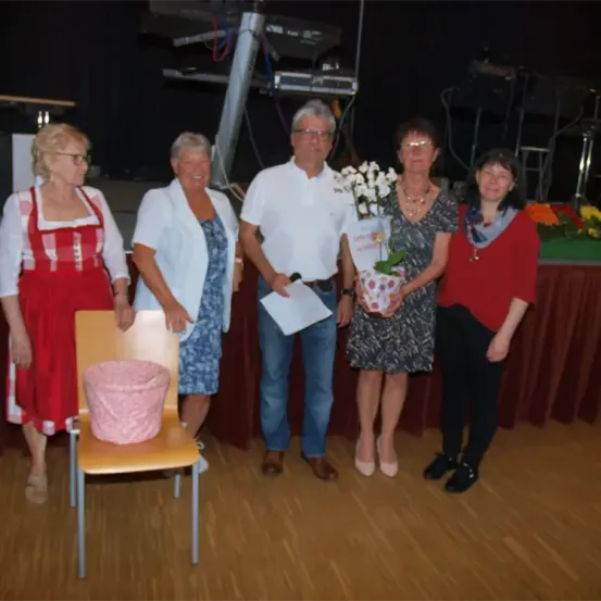 Fünf Personen stehen zusammen auf einer Bühne mit einem Tisch dahinter. Eine Person hält einen Blumenstrauß. Ein Stuhl mit einem rosa Korb steht neben ihnen.
