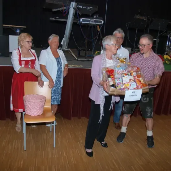 Fünf Personen stehen in einem Raum mit einer Bühne. Eine Frau in einem lilafarbenen Shirt hält einen Korb mit Geschenken. Ein Mann in einem karierten Hemd steht neben ihr. Dahinter steht eine weitere Frau in einem roten Kleid in der Nähe eines Stuhls mit einem rosafarbenen Korb.