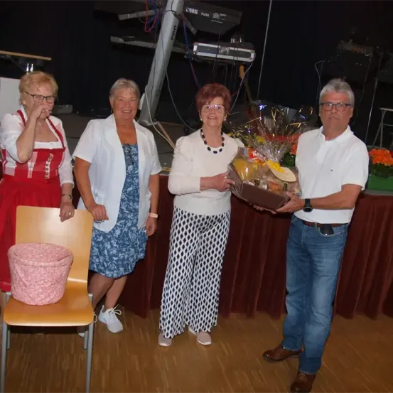 Vier Erwachsene stehen vor einem Tisch, eine Frau hält einen Korb. Der Mann im weißen Hemd hält einen Korb und die Frau im roten Kleid sitzt auf einem Stuhl.