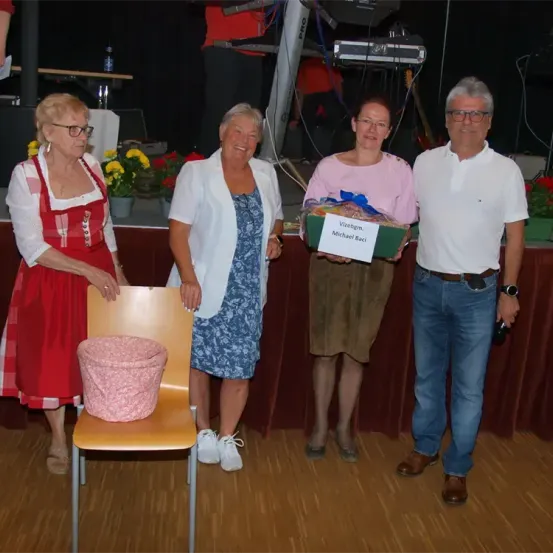 Vier Menschen stehen zusammen, möglicherweise bei einer Preisverleihung. Die Frau links trägt ein rotes Kleid. Die Frau in der Mitte hält eine Geschenkbox mit dem Text Vizebgm. Michael Baci.
