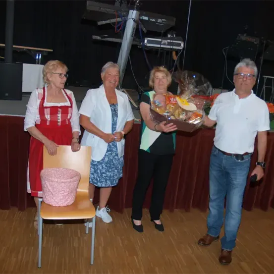 Vier Menschen stehen auf einer Bühne, eine Frau hält einen Geschenkkorb. Eine Frau trägt ein rotes Schürzen, eine andere einen weißen Blazer. Die Frau in Grün hält den Korb. Ein Mann in Jeans und einem weißen Hemd steht neben ihnen.