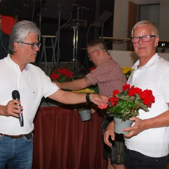 Zwei Männer in weißen Hemden halten Blumen und ein Mikrofon. Ein Mann in einem karierten Hemd sitzt hinter ihnen, mit einem Tisch und weiteren Blumen im Hintergrund.