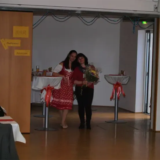 Zwei Frauen stehen neben Tischen, eine hält einen Blumenstrauß. Die Umgebung beinhaltet ein Bar-Schild und Dekorationen. Der Raum hat einen Holzboden und eine offene Tür.