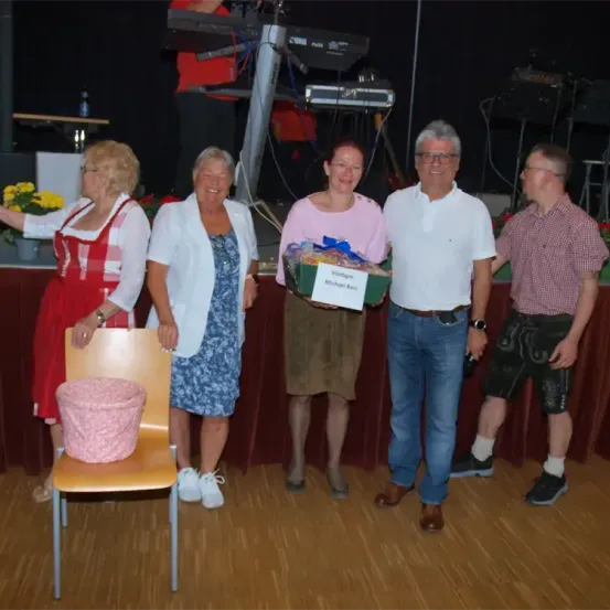 Fünf Menschen stehen in einem Raum mit Bühne und Musikinstrumenten. Eine Frau hält eine Geschenkbox und einen Korb. Eine weitere Frau lächelt neben einem Stuhl.