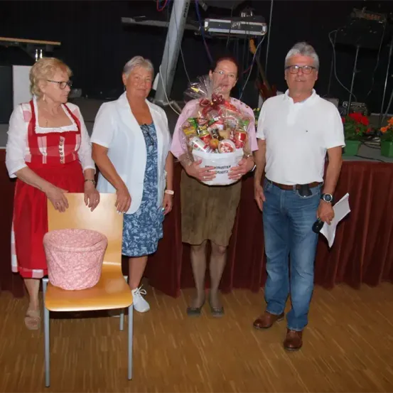 Vier Personen stehen gemeinsam auf einer Bühne. Zwei Frauen in traditioneller Kleidung, eine hält einen Stuhl mit einem rosa Kissen, die andere in einer weißen Jacke. Ein Mann hält einen Korb mit Geschenken, während ein vierter Mann in einem weißen Poloshirt und Jeans neben ihnen steht.