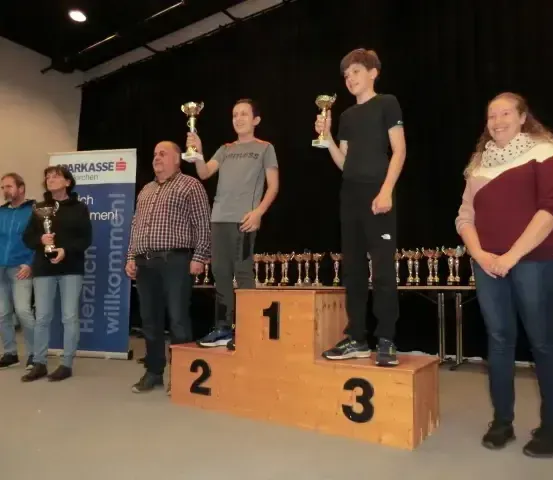 Die Gewinner stehen auf einem Podest mit Trophäen, wobei zwei Männer in der Mitte und eine Frau rechts stehen, während andere dahinter stehen.