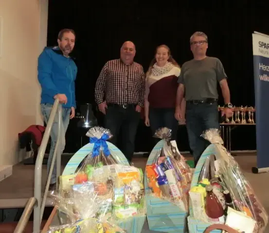 Vier Personen stehen hinter drei Geschenkkörben voller Lebensmittel und Weinflaschen, vor einem Banner, das Sparkasse Neukirchen sagt.