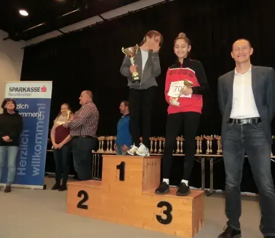 Drei Personen stehen auf einem Podest mit zwei Pokalen in der Hand, eine in der Mitte trägt ein rotes Shirt, und zwei weitere stehen dahinter. Ein Tisch mit Pokalen befindet sich rechts.