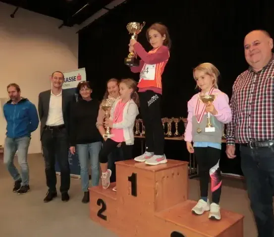 Ein junges Mädchen steht auf dem Podest des ersten Platzes, hält eine Trophäe und ist von anderen Kindern mit Medaillen und Erwachsenen umgeben. Sie befinden sich in einem Raum mit schwarzem Hintergrund und einer Fahne hinter ihnen.