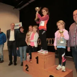 Ein junges Mädchen steht auf dem Podest des ersten Platzes, hält eine Trophäe und ist von anderen Kindern mit Medaillen und Erwachsenen umgeben. Sie befinden sich in einem Raum mit schwarzem Hintergrund und einer Fahne hinter ihnen.