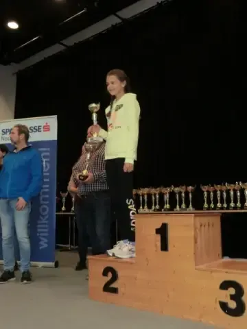 Eine Frau steht auf einem Podest und hält eine Trophäe in der Hand, mit zwei Männern hinter ihr. Das Podest ist von 1 bis 3 nummeriert. Der Hintergrund zeigt einen schwarzen Vorhang und ein Banner mit dem Text 'willkommen'.