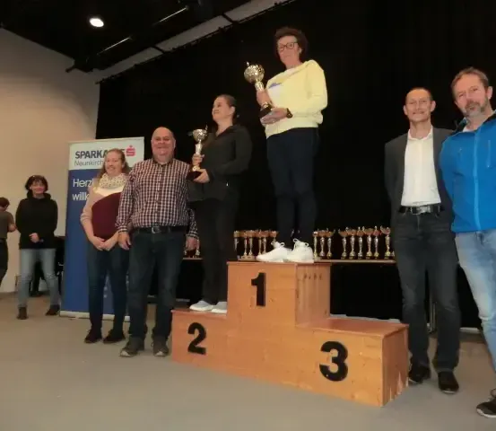 Eine Gruppe von Menschen steht auf einem Podest, eine Frau steht oben und hält eine Trophäe, die anderen halten ebenfalls Trophäen. Dahinter ist ein Banner zu sehen, und ein schwarzer Vorhang ist im Hintergrund.