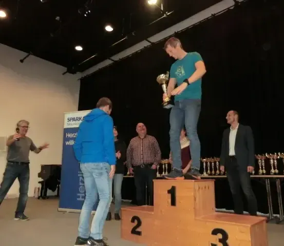 Ein Mann steht lächelnd auf einem Podest und hält eine Trophäe. Hinter ihm schauen Leute zu, und zwei Tische mit Trophäen sind vorhanden. Ein Klavier und ein Banner mit dem Wort 'Herz' sind sichtbar.