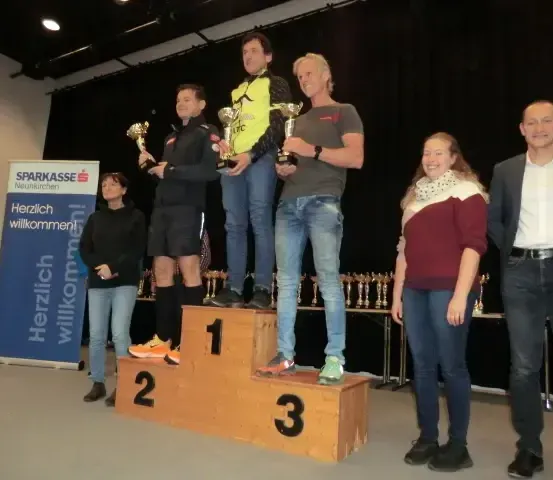 Die Gewinner stehen auf dem Podium, drei Männer halten Pokale und einer steht auf dem ersten Platz. Dahinter steht ein Banner mit der Aufschrift Herzlich willkommen.