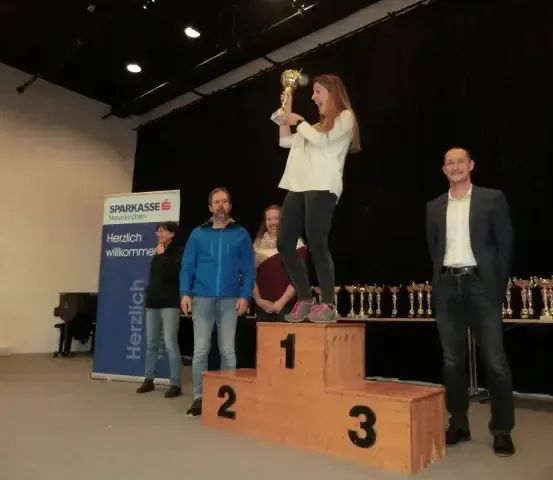 Eine Frau steht auf einem Podium und hält eine Trophäe in der Hand, während ein Mann auf der rechten Seite steht und einige Leute hinter dem Podium stehen, mit Trophäen auf dem Tisch.