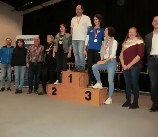 Eine Gruppe von Menschen steht auf einem Podium. Eine Person steht in der Mitte und zwei Personen sitzen an den Seiten. Alle tragen Medaillen. Sie lächeln und posieren für ein Foto. Hinter ihnen ist ein schwarzer Vorhang mit einem Banner.