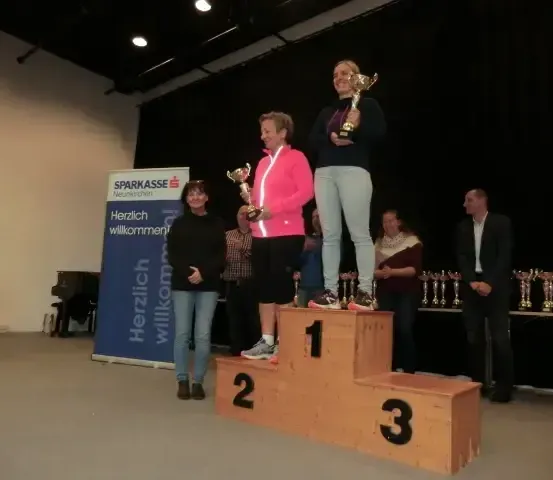 Drei Frauen stehen auf einem Podium mit einem blauen Hintergrund, auf dem Herzlich willkommen steht. Sie halten Trophäen und stehen auf den Zahlen 1, 2 und 3. Ein weiterer Mann steht hinter ihnen.
